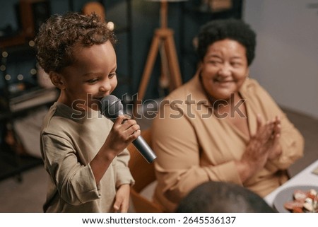 Child singing into microphone while elderly woman clapping hands with joy supporting cheerful grandchild during family gathering at home