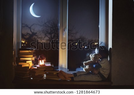 The child sits at night on the windowsill, hugs a toy and looks at the young moon.
