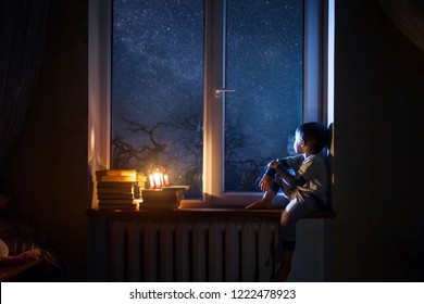 
The child sits on the windowsill at night looking at the stars and dreams. - Shutterstock ID 1222478923
