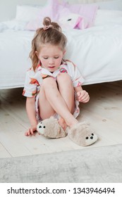 The Child Sits In Plush Slippers On The Floor In His Room