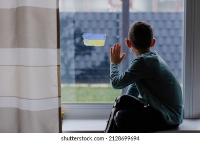 the child sits at the window with the flag of ukraine. State support in war. United Europe against Russia. Hope for the world. Anxiety and anticipation. Children's tears. Save Ukraine