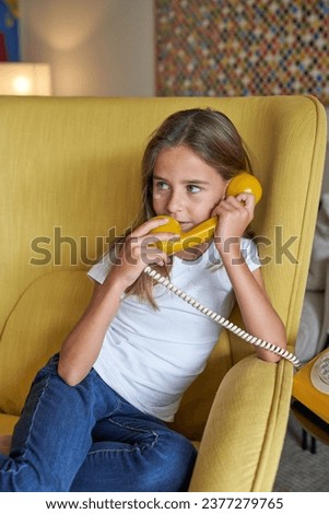 Child sitting in armchair in living room and talking on vintage rotary dial telephone while looking away at home