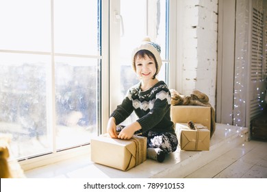 child is sitting by the window on a sunny Christmas day and makes out with gifts in boxes wrapped in paper.dressed in knitted warm woolen clothes and a hat. Inside the house with a stylish interior.