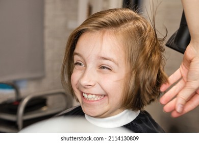 child sitting in hairdresser chair delightedly rejoices at new bob haircut. Hairdresser dries wet hair after cutting with hairdryer. Hair styling process in barbershop. Happy little girl