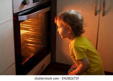 A Child Is Sitting Near The Oven In The Kitchen And Waiting. Curious Boy Is Watching Through The Glass Of Kitchen Oven. Baking Pizza, Muffins , Cupcakes Or Cookies.