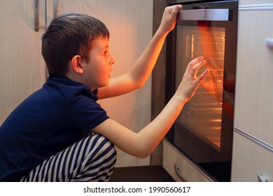 A Child Is Sitting Near The Oven In The Kitchen And Waiting. Curious Boy Is Watching Through The Glass Of Kitchen Oven. Baking Pizza, Muffins, Cupcakes Or Cookies.