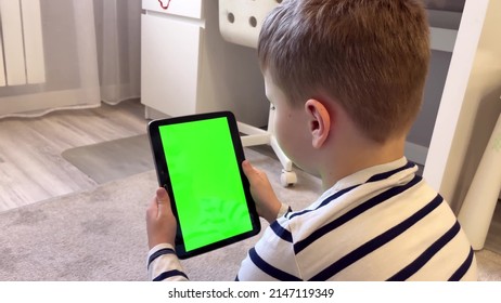 Child Is Sitting On The Carpet And Using A Digital Tablet PC With Green Screen, Back View. Boy Holding A Tablet With Green Screen In Hand