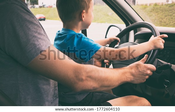 The child is sitting on the father\'s\
knees in the car and learning how to drive the\
car