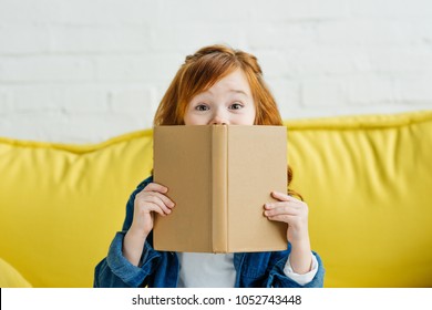 Child Sitting On Sofa And Holding Book In Front Of Her Face