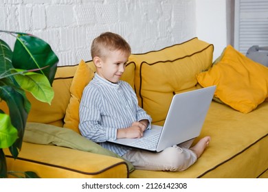 The Child Is Sitting On A Yellow Sofa With A Laptop. Carefully Looks At The Monitor.