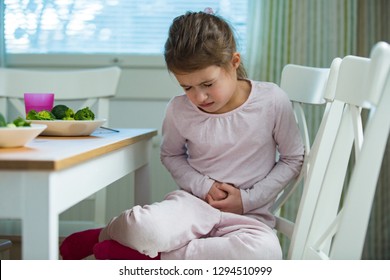 Child Sitting At The Table In The Kitchen With Stomach Pain. Hands On Belly. Little Girl Suffering