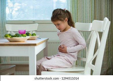 Child Sitting At The Table In The Kitchen With Stomach Pain. Hands On Belly. Little Girl Suffering