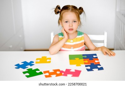 The child is sitting at the table with a sad look and looks at the camera questioningly. The girl can't solve the puzzle. Preschool development and education - Shutterstock ID 2258700021