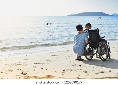 A Child Sitting In A Wheelchair Hugging His Father's Neck At The Seaside.