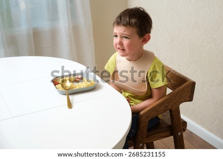 Child sitting at white table looking unhappy. Upset boy wearing bib and refusing to eat. Problem eating concept. A child aged 4 years (four years old)