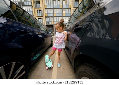 A Child With A Skateboard Stands Between Two Cars In A City Parking Lot. The Danger Of Skateboarding On Urban Streets And Roadways. Girl In Pink Shorts Learning To Ride A Skate Board