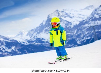 Child Skiing In Mountains. Active Toddler Kid With Safety Helmet, Goggles And Poles. Ski Race For Young Children. Winter Sport For Family. Kids Ski Lesson In Alpine School. Little Skier Racing In Snow