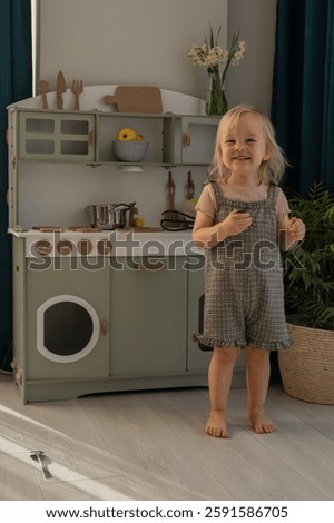 Child smiles while playing with kitchen toys in a well-decorated, light-filled room. Miniature kitchen features a variety of utensils and colorful items adding charm. Eco-friendly and safe handmade