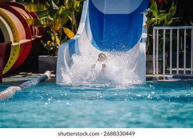 A child splashes into the pool at the end of a blue and white water slide at a tropical water park, surrounded by lush plants and colorful tube slides.
