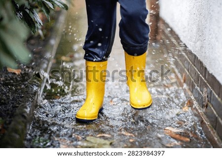 Child splashing through a flooded path in yellow rain boots and a raincoat, enjoying outdoor play during wet weather after heavy rain.