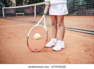 Child In Sportwear Playing Tennis On Outdoor Court. Cropped Image Of Child Legs On Tennis Court. Close Up View Of Tennis Ball, Racket And Shoes. Active Exercise For Kids.