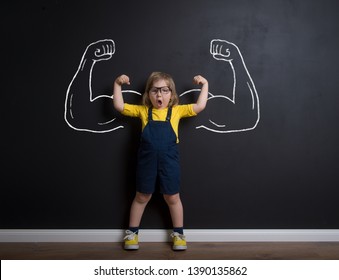 Child standing near chalkboard with muscles on it. Happy superhero kid is ready for kindergarten or daycare. Success, motivation concept. Girl power and feminism concept. Back to school.