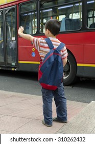 Child Standing On A Sidewalk Or Pavement Waving To A Red Public Transport Bus