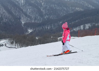A child stands on the skis on the ski slope. In the background is a winter forest with gray trees. Little skier at the ski resort