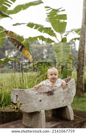 The child stands and smiles in a wooden cradle in the form of a boat against the backdrop of tropical greenery and palm trees