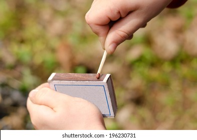 Child Striking A Safety Match On A Box Of Matches Against A Green Grass Background , Close Up Of The Hand