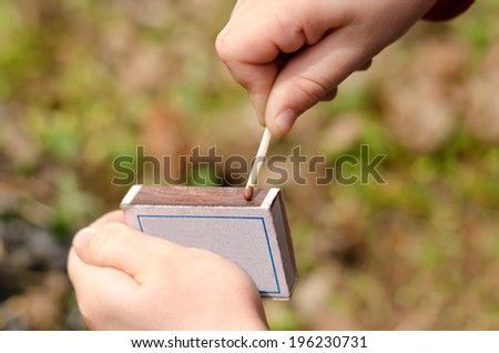 Child striking a safety match on a box of matches against a green grass background , close up of the hand