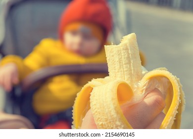 A child in a stroller eats a banana from his mother's hands, feeding children during a walk, a children's diet, a bitten fruit on a blurred background with a bokeh effect