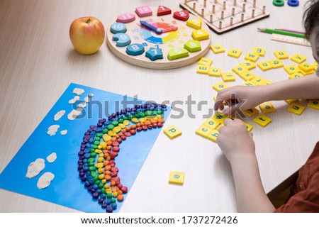 Child is studying, playing at home. Kid puts words from letters. Only hands are visible. At the desk art rainbow, clocks. Homeschool concept, distance learning, activity on self-isolation. Soft focus.