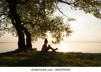 A child at sunset sits under a tree. Beautiful artistic photo with a kid girl in nature on a sunny summer evening. Dreams, fantasies, inspiration, childhood, inner world and child's life concept.