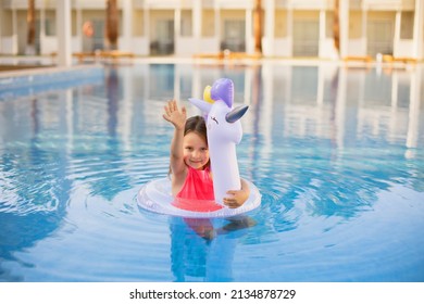 Child In Swimming Pool Waving Her Hand . Little Girl Playing In Water. Vacation And Traveling With Kids. Children Play Outdoors In Summer. Kid With Unicorn Inflatable Ring Toy.