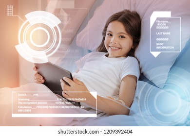 Child With Tablet. Cheerful Emotional Girl Recovering From Illness In Hospital And Smiling While Staying In Bed With A Modern Tablet In Her Hands