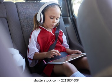 Child With Tablet, Headphones And Safety Belt In The Car Going Home After Soccer Match, Game Or Practice. Young Girl In Sports Gear Playing Educational Games Online On Tech Device After Training