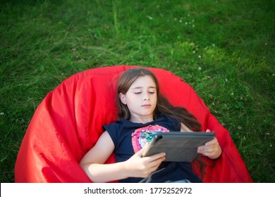 Child With Tablet In His Hands Is Lying On Grass. Girl Looks At Her IPad In Summer On Lawn. Person Reads And Learns.