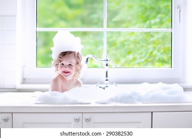 Child Taking Bath. Little Baby In A Kitchen Sink Washing Hair With Shampoo And Soap. Kids Playing With Foam And Water Splashes. White Bathroom With Window. Clean Kid After Shower. Children Hygiene. 