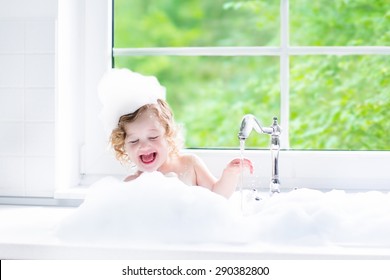 Child Taking Bath. Little Baby In A Bath Tub Washing Hair With Shampoo And Soap. Kids Playing With Foam And Water Splashes. White Bathroom With Window. Clean Kid After Shower. Children Hygiene. 