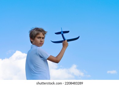 Child Throwing Toy Plane. Kid Holding Plane And Looking Direct To The Camera. Boy Playing With Blue Toy Airplane Against Blue Sky On Sunny Day Outdoors