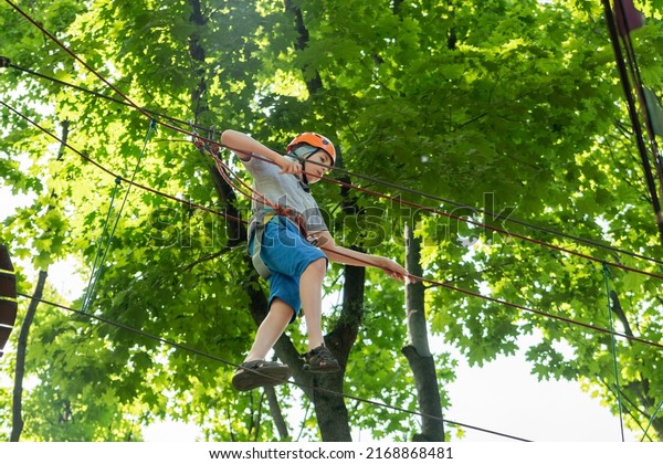 The child is a tightrope walker. Rope park. A boy\
teenager in a helmet walks on suspended rope ladders. Carabiners\
and safety straps. Safety. Summer activity. Sport. Children\'s\
playground in nature