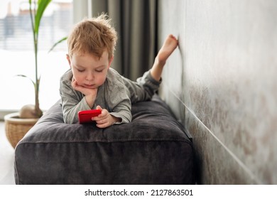 Child, Toddler Playing With Smartphone, Lying On The Sofa At Home. Screen Time Limit - Concentrated, Involved Little Boy Looking To Mobile Phone.