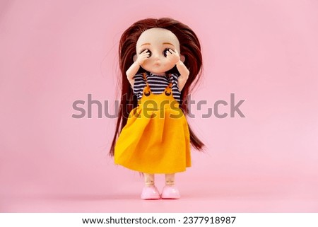 A child toy doll with dark hair in an orange dress standing on a pink background. Plastic children's toy. Doll games for imagination. the frightened doll closes her eyes in fear.