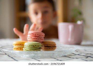 Child trying to catch a stack of delicious macaroons, of three colors, on a table with a checkered tablecloth, and a cup of coffee in the background