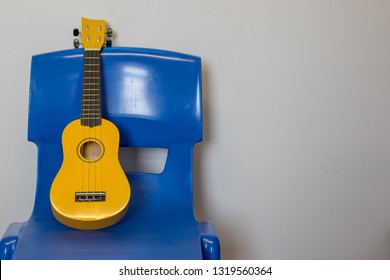 Child Ukulele On Chair. School Music Lesson Practice Room. Bright Yellow Uke On Blue Plastic Chair. Childrens Musical Instrument.