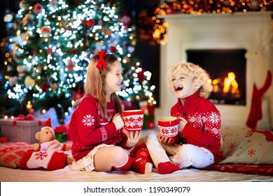 Child Under Christmas Tree At Home. Little Boy And Girl In Knitted Sweater With Xmas Ornament Drink Hot Chocolate. Family With Kids Celebrate Winter Holidays. Kids Open Presents At Fireplace.