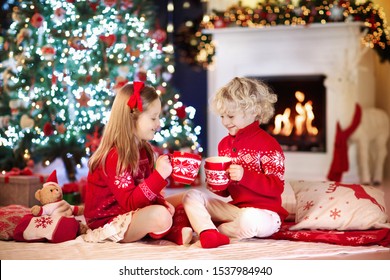 Child Under Christmas Tree At Home. Little Boy And Girl In Knitted Sweater With Xmas Ornament Drink Hot Chocolate. Family With Kids Celebrate Winter Holidays. Kids Open Presents At Fireplace.