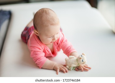 A Child Under One Year Old In A Pink Suit Lies On His Tummy On A White Sofa And Plays With A White Cat Toy