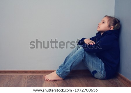 A child upset, oppressed, offended, in despair sits on the floor in the corner against the background of a gray wall. A little girl has problems with her parents, with friends, with peers, at school